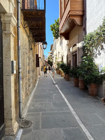 The image depicts a narrow, stone-paved alleyway in Rethymno, Greece, bathed in sunlight. The buildings flanking the alley are constructed of weathered stone and plaster, with wooden balconies and ornate architectural details. The left side of the alley features a rough-textured stone wall with a downspout and an intercom panel. The right side of the alley is lined with large terracotta pots containing lush green plants. Several people are walking away from the viewer down the alley, dressed in casual summer clothing. The ground is paved with large, irregular stone slabs with a white line painted down the center of the alley. The sky is clear blue.