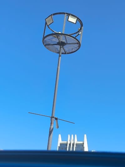 The image displays a clear blue sky background with a prominent, tall metal utility pole. At the top of the pole, a circular high-mast lighting structure is mounted. This structure consists of a lower circular mesh platform and an upper metal ring, within which at least three square-shaped LED floodlights are visibly affixed. A small, yellowish spherical object is positioned centrally within the lower section of this lighting array. Further down the pole, a horizontal metal bar with two shorter, outward-extending arms is clamped to the pole. In the lower right background, a portion of a modern white building with distinctive vertical architectural elements and dark windows is visible. A blurred, dark blue horizontal element occupies the bottom of the frame, suggesting a foreground obstruction. No individuals are present. The scene is located in Kızılbaş, Northern Cyprus / Cyprus.