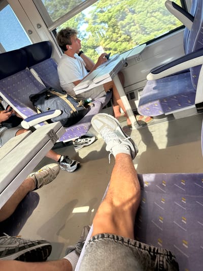 The image depicts the interior of a train carriage with an emphasis on the seating area and passengers' legs and feet. A young man wearing a white t-shirt and shorts is seated by the window, looking out. He has earbuds in and appears to be holding a phone or similar device. The seats are upholstered in a fabric with a purple base and a pattern of white and yellow lines. A backpack is placed on a seat next to the man. Several pairs of legs and feet, clad in various athletic shoes, are visible throughout the frame, suggesting the presence of multiple passengers. The train is in motion, as indicated by the trees and greenery visible outside the window, suggesting the train is traveling through a rural or suburban area, likely in or near Salses-le-Château, France. The lighting suggests it is daytime, with sunlight filtering into the carriage.