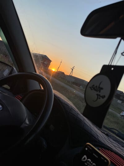 The image provides an interior view from a car, looking out the windshield at a sunset. The scene is set in a rural or semi-rural area, with buildings and power lines visible against a soft orange and blue sky. The sun is low on the horizon, casting a warm glow.
Dominant objects within the car include the steering wheel, a rearview mirror, and a dashboard. A patterned car seat cover is visible on the passenger side. Hanging from the rearview mirror is an air freshener with an oval tag that has some script written on it, appearing to be in Arabic or a similar script. A smartphone is also visible on the dashboard, showing a lock screen with some text.
The time of day is clearly sunset, given the low angle of the sun and the warm, diffused light. The weather appears to be clear. The location cues, such as the buildings and power lines, suggest an outdoor setting in a developed, though possibly sparse, area. There are no people visible in the image. The overall mood is serene and reflective, perhaps conveying a sense of journey or transition associated with the end of the day.