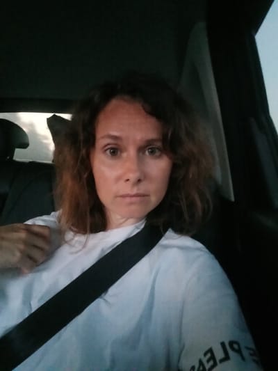 A woman with auburn, wavy hair is seated in the driver's seat of a car, wearing a white t-shirt and a black seatbelt. She is looking directly at the camera with a neutral expression. The interior of the car is dark, with the sky visible through the rear window, suggesting it is either dawn or dusk. The image is a selfie, taken from a slightly high angle. The location is identified as Sultaeva, Russia.