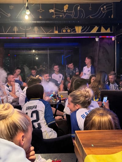 In Travnik, Bosnia and Herzegovina, a group of people are gathered indoors, likely at a cafe or lounge, marked by ambient lighting and wall art. The scene is filled with cigarette smoke, suggesting a social gathering where smoking is permitted. Several individuals are seated, engaged in conversations or watching something, with one person in the foreground smoking a shisha pipe. A woman in the foreground is holding a smartphone, potentially capturing the moment. The environment appears casual and dimly lit, with several individuals wearing athletic-style jerseys, some with names and numbers visible on the back, hinting at a shared interest or team affiliation.