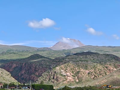 The image shows a mountainous landscape under a bright blue sky with scattered clouds. In the foreground, there are rolling hills covered with sparse vegetation and rocky outcrops. A deep gorge with steep, reddish-brown rock faces is visible in the mid-ground. In the far distance, a large mountain peak is shrouded in mist, with some snow visible at its summit. At the very bottom of the image, there are some signs of human activity, including what appears to be a construction site with an excavator, a few vehicles, and some low-lying buildings partially obscured by vegetation. The scene depicts a rural or remote area, likely outside of the main urban center of Cholpon Ata, Kyrgyzstan, given the natural landscape. The overall impression is one of a vast, rugged natural environment with some ongoing development.