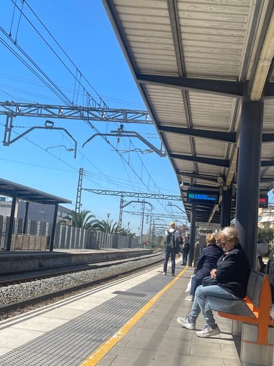 The image captures a daytime scene at a train station platform. Overhead, a metal structure provides shelter. The sky is clear and blue. Power lines and related infrastructure crisscross above.

People are present on the platform, including a couple seated on orange seats and a few others standing or walking.

The platform edge is marked with a yellow line. The train tracks are visible alongside the platform, running parallel with a gravel-covered path between the tracks.

A digital display indicates "R2 Barcelona - Estacio de França" in blue text. Based on this, the location is Gavà, Spain, part of the Barcelona metropolitan area, as this is a train line connecting Gavà to Barcelona's Estació de França. Other buildings and trees can be seen in the background.