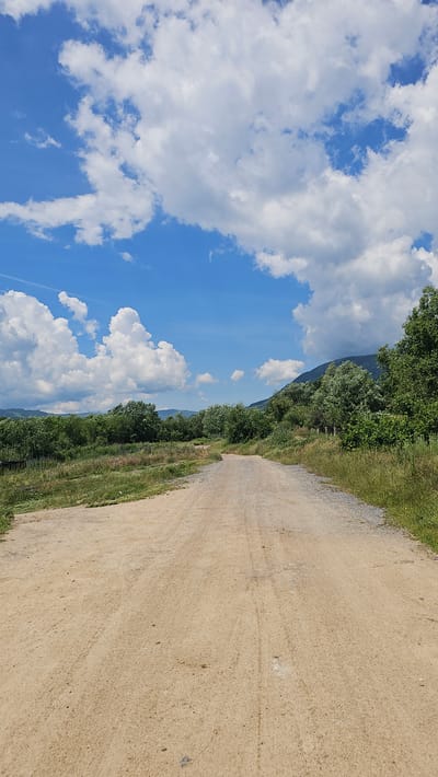 A dirt road curves through a rural landscape under a bright blue sky dotted with fluffy white clouds. The road, composed of light brown dirt and gravel, stretches into the distance, flanked by green vegetation, trees, and rolling hills. The scene appears to be a daytime view in the village of Rozino, Bulgaria.