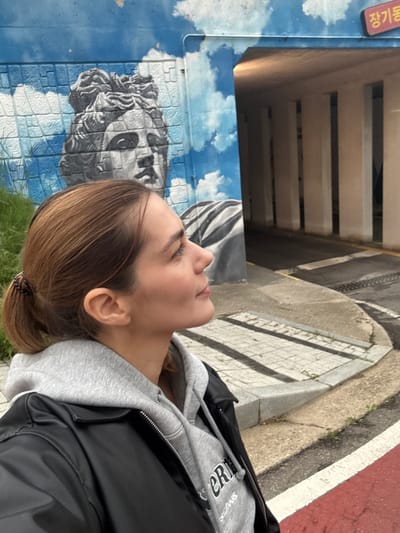 A woman stands next to a mural of a classical sculpture on a blue brick wall, likely in Gimpo-si, South Korea. She is wearing a grey hooded sweatshirt under a black jacket. The environment includes what appears to be a sidewalk, a building facade with wooden slats, and a street. The mural has a cloudy sky background.