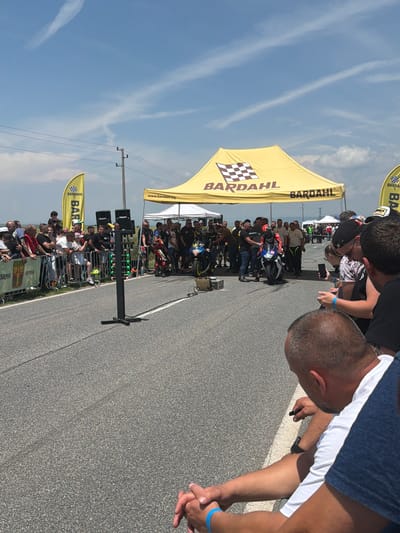 An outdoor scene captures a motorcycle drag racing event occurring on a paved asphalt road in Chirpan, Bulgaria. The sky overhead is clear blue with multiple white contrails visible.
In the immediate foreground, the backs of several spectators' heads and shoulders are visible, indicating their close proximity to the event. A male spectator in a white t-shirt, wearing a blue wristband on his left wrist, is seated on the ground with his hands clasped, observing the scene to the left.
The midground features the race starting area. A black "Christmas tree" starting light system stands prominently on the left side of the asphalt track, equipped with multiple green and red lights. Two large black speakers are mounted atop this structure. Electrical cables extend from the base of the starting system across the road surface.
To the right of the starting system, a motorcyclist in a full red, black, and white racing suit and helmet is positioned on a white and blue sport motorcycle, ready for the race. Another individual stands near this motorcycle. Further to the left and slightly behind, a second motorcyclist in a blue, black, and white racing suit and helmet is visible on a blue and white sport motorcycle.
A crowd of spectators is gathered behind metal barriers lining both sides of the road. On the left, a fence banner displays the text "ОБЩИНА ЧИРПАН" (Municipality of Chirpan) along with a crest, confirming the event's location.
In the background, a large yellow tent with the "BARDAHL" logo and a checkered flag is erected, spanning across the scene. The text "motor oils and additives" is partially visible beneath the logo. The side of the tent also displays "www.bardahl.bg". To the left of the main tent, a yellow banner flag with the "BARDAHL" logo is also present. A smaller white tent is visible behind the main yellow tent. Utility poles and power lines traverse the distant horizon, which features a flat, light-green landscape under a hazy sky.