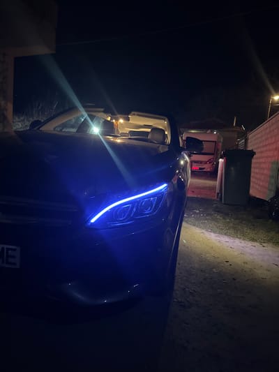The scene depicts a nighttime outdoor environment in Bohot, Bulgaria. A dark-colored convertible vehicle, possibly a sedan or coupe with its roof retracted, occupies the foreground, parked on a dirt or gravel surface. The front left quarter of the vehicle is visible, featuring a prominent blue LED daytime running light illuminated on the left headlight assembly. A partially visible Bulgarian license plate on the front bumper displays the letters "ME." The car's interior shows light-colored seats and a bright internal light source, causing a lens flare near the windshield.

In the mid-ground, a white motorhome branded "HYMER" is parked, facing towards the viewer. To the right of the motorhome and positioned along a brick wall, a dark green wheelie bin with a black lid is visible. The brick wall exhibits a reddish tint on its upper section. A street light or similar outdoor luminaire is present in the upper right, casting a yellow-orange glow. The ground immediately to the right of the car is illuminated by a localized yellow-toned light beam. No individuals are present in the image.