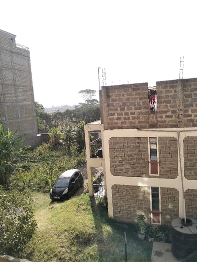 A residential scene shows a black car parked in a grassy area next to a two-story, cinderblock building. The building is under construction, with the top floor still exposed brick and rebar. Laundry hangs in an unfinished window. Nearby is a water tank. In the background are trees and another multi-story, cinderblock building, appearing incomplete. The sky is bright and hazy.