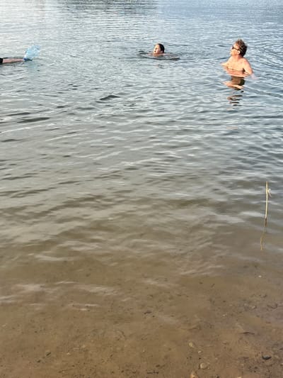 In Zamost'e, Russia, on a body of water, a group of people are engaged in swimming. A person on the left is holding a large plastic water bottle. In the mid-ground, a child is swimming, and to their right, an adult man with sunglasses is also in the water, looking upwards. The water is calm with visible ripples, and the bottom is sandy with scattered pebbles. A thin reed or stick is visible sticking out of the water on the right side of the image. The scene depicts a casual recreational activity in a natural setting.