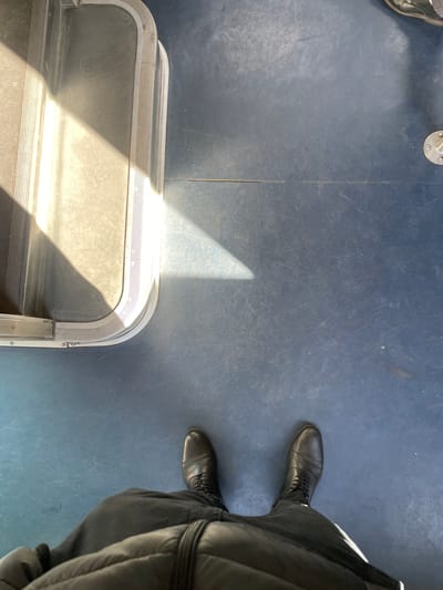 The image displays a top-down, first-person perspective within a public transport carriage, likely a train or tram, in the vicinity of Saint-Cyr-l'École, France. The individual's lower body is visible, wearing dark trousers and black, lace-up leather boots. A portion of a dark, possibly quilted jacket is present at the very bottom edge. The floor is a worn, dark blue or gray-blue linoleum surface, exhibiting numerous scratches and specks of dust. To the left, a metallic threshold or step frame with distinct ridged anti-slip strips is illuminated by a strong, trapezoidal beam of sunlight, which also casts a sharp shadow diagonally across the floor. A partial view of a circular metallic fixture, fastened by screws, is visible on the upper right side of the floor.
