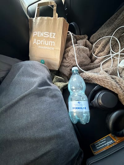 The image shows the interior of a vehicle. In the foreground, a person's dark denim-clad legs are visible. To the right of the legs, a brown paper bag with "PENSEZ Aprium PHARMACIE" printed in white text is situated. Next to the bag is a bottle of "EGEKILDE" natural mineral water. Further to the right, a pair of black Sony headphones rests on a brown, textured blanket. A white cord is also visible draped over the blanket. In the bottom right corner, a package labeled "Acrylic Paints Acrylfarben Peinture acrylique" is present. The overall scene suggests items belonging to a passenger in the vehicle. The location is identified as Ödsmål, Sweden.