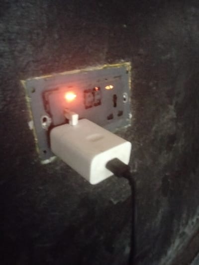 The image captures a close-up view of an electrical wall socket with a charger plugged into it, set against a dark, textured background.

The central focus is a rectangular electrical outlet panel, likely made of grey or dark-colored plastic, which is recessed into the wall. Around the perimeter of the socket panel where it meets the wall, there is a visible line of yellowish or light-colored material, possibly grout or sealant, indicating its installation.

One of the switches on the left side of the socket panel is illuminated with a prominent, bright orange-red glow, suggesting that the socket is currently switched on and receiving power. To the right of this illuminated switch, several other slots and potential switches or indicators are visible, though less distinctly. The socket appears to be a multi-standard type, capable of accommodating different plug configurations.

A white, rectangular power adapter (charger) is plugged into one of the outlets on the panel. A black cable is plugged into the bottom end of this charger and extends downwards out of the frame.

The wall itself is very dark, appearing black or very dark grey, and has a visibly rough, textured surface. The ambient lighting in the scene appears dim, making the glow from the socket switch particularly noticeable. No people, discernible text, or specific outdoor elements are visible in the image. The activity depicted is the use of an electrical outlet to power a charging device.