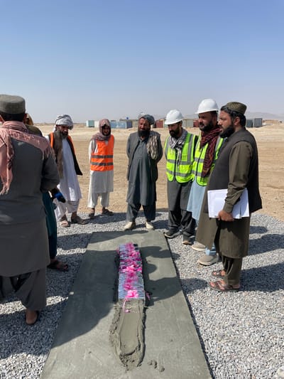 In Pir Dust, Afghanistan, ten adult males are gathered around a freshly poured concrete slab on a dry, gravel-covered ground. The men are attired in traditional Afghan clothing, with four wearing safety vests (three lime green, one orange) and two wearing white hard hats. The man on the far right holds a stack of white papers.
At the center of the gathering, a rectangular object, approximately 70 centimeters long, is partially set into wet concrete within a channel on the slab. This object is wrapped in a reflective material, festooned with pink flowers, and covered by a layer of clear plastic sheeting. The immediate surroundings consist of light grey gravel. In the background, a vast, arid, and dusty plain extends to the horizon, dotted with several light-colored shipping containers or modular units, with distant mountains visible beneath a clear blue sky. Strong shadows indicate bright, direct sunlight.