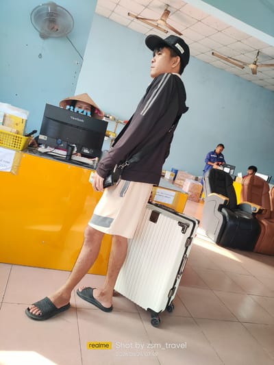 A man stands with a white suitcase at a counter in what appears to be a post office or shipping facility. He is wearing a black baseball cap, a black jacket with white stripes, and light-colored shorts. His right foot is on a black slide sandal, and his left foot is also in a black slide sandal. The counter is bright yellow, and behind it, a person wearing a conical hat is partially visible looking at a computer monitor. To the right of the counter, there are several large reclining chairs, one black and one brown, and two men in blue uniforms are standing near a stack of boxes. In the background, there are more boxes and some ceiling fans. The scene is lit by bright artificial light. The time and date stamped on the image are 2026.06.24 07:49. The visible text includes "KẾ TOÁN 5575", "ViewSonic", "DAFI : HUỆ NGHĨA GD SG MẬT KHẨU, 19006471", and "realme Shot by zsm_travel". The location context provided is Gò Dầu, Vietnam.