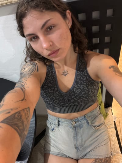 This image, rotated 90 degrees counter-clockwise from its original orientation, features a young woman taking a close-up selfie indoors.
The subject is a woman with long, dark, wavy hair. She has a neutral expression, looking directly at the camera. Her face shows some freckles or blemishes, and she has piercings in her earlobes and a ring in her nose. She is dressed in a grey and black patterned sports bra, featuring what appears to be an animalistic or leopard print, and light blue denim shorts.
She has multiple tattoos: a small, stylized sun symbol on her chest, just below her collarbone, and extensive black and grey sleeve tattoos covering large areas of both arms with intricate, possibly abstract or nature-inspired designs.
The setting appears to be a casual indoor space. The woman is seated or lounging on dark furniture with a backrest featuring square cutouts. Behind her, a green pillow and another pillow with a light-colored, patterned cover (reminiscent of a mandala design) are visible. The background includes a light-colored wall and a light brown or beige tiled floor. A dark table or surface is partially visible in the foreground on the right.
The activity is a self-portrait, indicated by the camera angle and the subject's direct gaze. The lighting is consistent with an indoor environment. No specific time of day or outdoor weather can be determined from the image. While the provided location context is Burj El Brajneh, Lebanon, there are no specific visual cues within the image itself to confirm this geographical detail. No readable text is visible in the photograph.