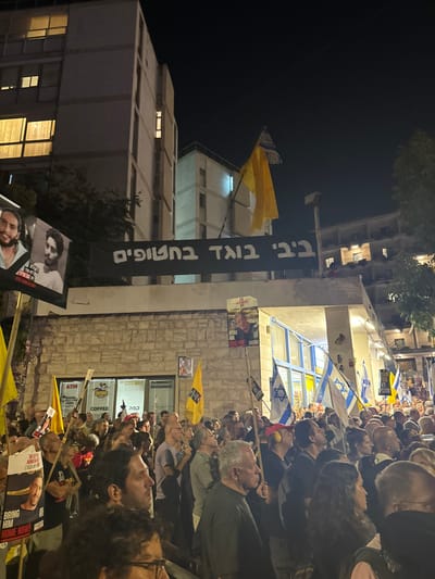 This image depicts a nighttime protest in an urban area of West Jerusalem, Israel. A large, dense crowd of individuals, primarily adults, occupies a street or plaza, gathered in front of multi-story buildings.

**People:** Hundreds of protesters are visible, densely packed, facing generally forward and slightly upwards. They are dressed in various casual attire.

**Objects and Subjects:**
*   **Banners and Signs:** A large black banner with white Hebrew text, "ביבי בוגד בחטופים" (Bibi betrays the hostages), is prominently displayed across the upper facade of a ground-level building. To its immediate left, another large banner features two photographic portraits of young men, likely related to the hostage crisis. Within the crowd, numerous handheld signs are visible; some display photographs of individuals and text such as "BRING HIM HOME NOW!", while at least one sign features a photograph of Benjamin Netanyahu.
*   **Flags:** Multiple Israeli national flags are held aloft by protesters. Several large, plain yellow flags, some with a black-and-white checkered pattern at the top, are also interspersed throughout the crowd. These yellow flags are commonly associated with the "Bring Them Home Now" movement.
*   **Buildings:** The background features several multi-story buildings. The building immediately behind the protesters has a stone-clad ground floor with visible storefront signs for an "ATM," "DODO COFFEE," and a "CURRENCY EXCHANGE." Above this, and extending further into the background, are concrete residential or commercial structures with illuminated windows, indicating occupancy.

**Actions and Interactions:** The individuals are gathered in a concentrated formation, holding signs and flags, indicating a public demonstration or protest. Their collective gaze suggests attention directed towards a focal point beyond the immediate foreground.

**Environment:** The scene is illuminated by artificial light sources from the buildings and streetlights, set against a dark night sky. The architecture signifies a typical urban setting.

**Location:** The event is situated in an urban commercial and residential district within West Jerusalem, Israel. The specific street is not identifiable from the visual information provided.