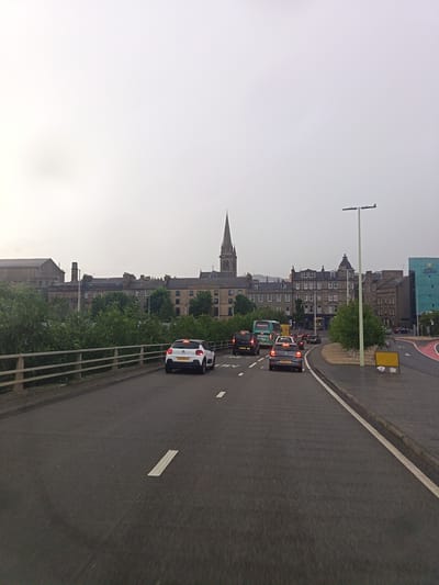 The image depicts a street scene in Dundee, United Kingdom, on an overcast day. The foreground shows a road with several cars in traffic. A white Citroen hatchback is visible in the middle of the frame, while other cars, including a black minivan and a silver Volkswagen hatchback, are ahead of it. A tour bus with "COOPER'S TOURMASTER" written on its side is also in the traffic.
In the background, a line of buildings forms the city skyline. A prominent spire, likely from a church or cathedral, rises above the rooftops. The architecture suggests an older European city. Green trees line the left side of the road, separated from the traffic by a wooden fence. A modern blue building with the "Express by Holiday Inn" logo is visible on the right side of the frame, indicating a commercial area. A lamppost stands tall on the right side of the road.
The overall atmosphere is mundane, with the traffic and overcast sky suggesting a typical day. The lighting is diffused, typical of an overcast sky, and there is no strong sunlight. The time of day is likely daytime, given the visibility and the activity on the road. The image is captured from a moving vehicle, giving a slightly blurred perspective of the road markings and the immediate surroundings. The license plates of the visible cars are CK17 EVJ1 and NN16 VEZ.