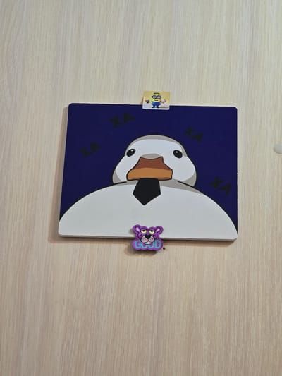 The image shows a square, dark blue canvas depicting a cartoon white duck with a black tie and orange beak. The duck's eyes are black. Scattered across the blue background are several black symbols that resemble the letter "X" or a stylized "A". Attached to the top edge of the canvas is a small square sticker featuring a yellow Minion character holding a cup. Below the canvas, attached to its lower edge, is a purple cartoon bear or cat-like creature with a bow and the word "GOOD" embroidered in blue lettering. The canvas and attachments are mounted on a light-colored wooden surface, likely a door or wall, with a visible wood grain pattern. The scene is indoors. The location in Novocherkassk, Russia is not discernible from the image.