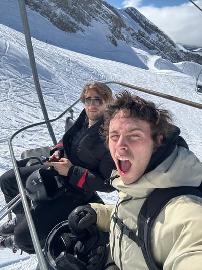 Two males are depicted riding a ski chairlift upwards through a snowy mountain landscape, consistent with a ski resort in La Giettaz, France. The subject in the foreground, a male with curly brown hair, wears a light beige ski jacket and a black backpack. He looks towards the camera with his mouth wide open and eyes squinted, holding a black ski helmet with gloved hands. Behind him, a second male with lighter hair and sunglasses is seated, wearing a dark puffer jacket with red accents. He holds a black smartphone in his right hand and has a black ski helmet resting on his lap. The background features extensive snow-covered ski slopes with visible tracks, leading up to steep, rocky mountain peaks under a partly cloudy sky. Metal elements of the chairlift frame are visible throughout the image.