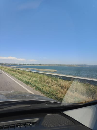 The image captures a wide, clear blue sky above a serene body of water, likely a lake or reservoir, stretching towards a distant, hazy horizon. The water's surface is calm, with subtle ripples reflecting the sunlight. Along the left side of the frame, a paved road with a white line marking its edge runs parallel to the water's edge. Beside the road, tall, dry grasses and reeds grow, interspersed with patches of greener vegetation. A concrete barrier separates the grassy embankment from the road. In the background, a low-lying town with numerous buildings is visible along the far shore of the water. The foreground of the image shows the dashboard and windshield of a vehicle, with the reflection of the grassy embankment and the sky visible on the windshield. The scene suggests a peaceful, rural or semi-rural setting, possibly along a coastal highway or a road bordering a large body of water. The location is identified as being in the city of Koshary, Ukraine.