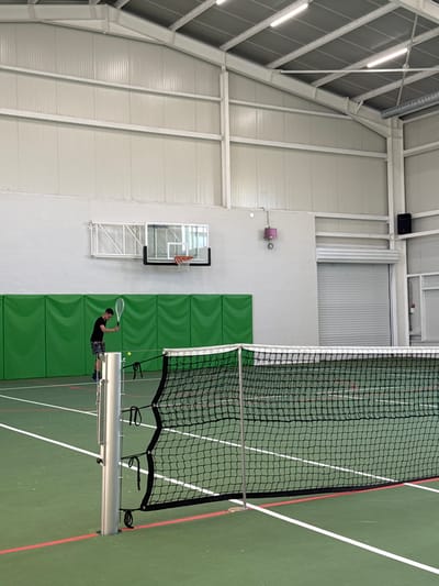 The scene depicts an indoor sports facility in Gorna Mitropolia, Bulgaria. A green-surfaced tennis court, marked with white and red lines, occupies the foreground and midground, bisected by a black tennis net with a white top band supported by silver metal posts. One male individual is present on the far side of the net, facing away from the camera. He wears a dark short-sleeved shirt and dark patterned shorts, holding a white tennis racket in his right hand. A yellow tennis ball is visible airborne in front of him, suggesting a serve or hit in progress. The background wall features a lower section of horizontal green padded panels, with the upper wall being a light color. A wall-mounted basketball hoop with a transparent backboard, bearing the text "inteplastic," and an orange rim is visible above the green padding. To the right of the hoop, a pink/purple box-like device is mounted, and further right, a large grey roll-down security door is closed. A black speaker is mounted high on the right wall. The ceiling features exposed metal trusses, corrugated panels, and linear fluorescent lighting fixtures.