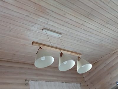 The image shows a wooden ceiling with a pendant light fixture. The fixture has a light-colored rectangular mounting plate on the ceiling from which three white conical lampshades hang. Each lampshade has a brown leather strap that attaches it to the fixture. The lampshades are arranged in a line and appear to be made of metal. The ceiling is constructed of light-colored wooden planks. The scene is viewed from below, looking up at the ceiling and the light fixture. There are no people or animals visible in the image. The location is identified as Sokyrna, Ukraine.