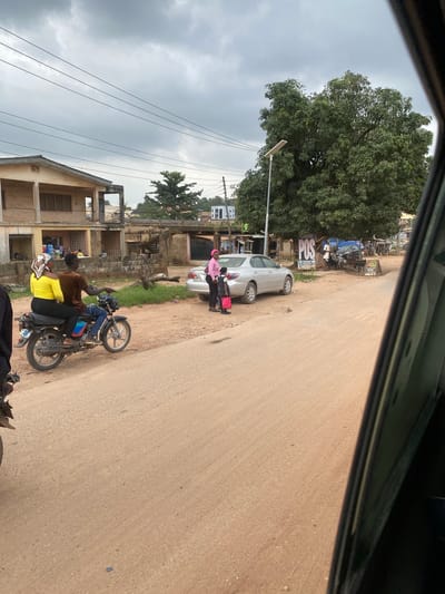 In the city of Ojere, Nigeria, an overcast sky looms over a scene of everyday life on a dirt road. A man and woman ride a motorcycle, the woman in a yellow top and the man in a brown shirt. They are positioned in the foreground, with the motorcycle angled towards the right of the frame. Further down the road, a silver sedan is parked. Near the car, a woman in a pink shirt and a child in a school uniform with a red bag stand on the roadside, possibly interacting with a nearby POS (Point of Sale) machine. Two other individuals are visible near a shelter with "POS" signage, suggesting a commercial activity. Buildings line the street, some appearing to be residential with shop fronts at ground level. A large tree with dense foliage dominates the mid-ground, and a streetlamp stands near it. The environment is dusty, with loose soil forming the road surface.