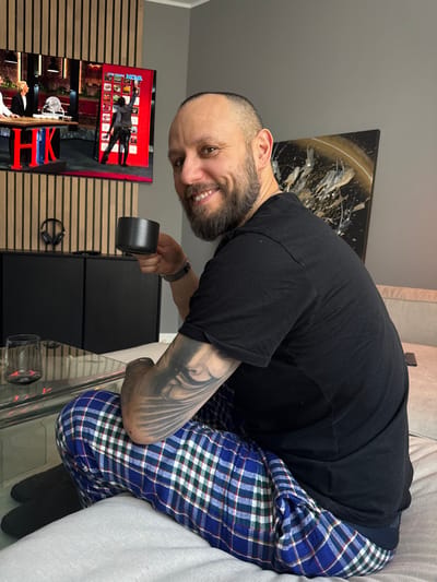 The scene depicts an indoor domestic environment, located in the city of Ålesund, Norway. An adult male with short hair and a beard is seated on a light-colored sofa, angled towards the right, with his head turned to face the camera, displaying a smile. He is wearing a black short-sleeved t-shirt and blue plaid pajama pants with green and white lines. A metallic watch is visible on his left wrist, and his left forearm displays a tattoo featuring a stylized facial profile with radiating lines. He holds a dark grey, ribbed ceramic mug in his right hand.

In the foreground, a clear glass coffee table features a clear glass containing a dark liquid. The background wall, directly behind the man, is painted grey. To the left of the man, a large flat-screen television is mounted on a wall section featuring vertical light-wood slats. The television displays a program: on the left side, two individuals, one in a white chef's coat, are visible next to a wooden counter with a red "HK" logo; on the right side, a person in dark clothing is shown reaching towards a red vending machine-like structure labeled "POLIGON NOVA". Beneath the television, a black two-door cabinet is positioned, with black over-ear headphones hanging on the wall above it. To the right of the television, on the grey wall, an abstract artwork is mounted, featuring a black background with white and gold splatters and a curved line.