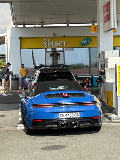 At a Shell gas station in Drazhevo, Bulgaria, a blue Porsche convertible is parked, facing away from the camera. The car has the license plate "CB 4403 OA" and "Porsche Zentrum Friedrichshafen" printed below it. Behind the Porsche is a black SUV with a roof rack. Two individuals are standing near the SUV, a woman in athletic wear on the left, and a man in a yellow t-shirt and denim shorts on the right. The gas station features a yellow "Shell Select" sign and a "Shell café" logo. Fuel pumps with hoses are visible to the right of the vehicles. The scene is outdoors under daylight.