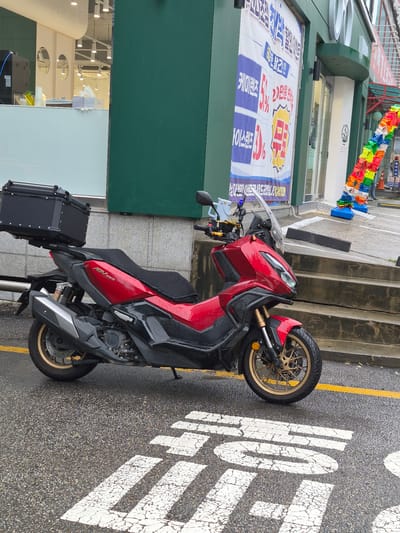 A red and black Honda ADV 350 scooter is parked on a wet, asphalt street in Suwon-si, South Korea. The scooter has a black storage box mounted on the rear and gold-colored wheels. In the background, a building with a green facade has a large banner with Korean text and graphics, and a string of colorful decorative elements hangs near the entrance. The street markings are white and partially obscured by water.