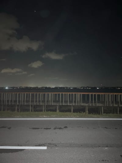 A night landscape shows a dark sky with scattered clouds. A body of water reflects the faint city lights on the horizon. A structure, possibly a pier or boardwalk, extends along the water's edge. A paved road runs parallel to the structure, marked with a white line. The overall scene is dark and slightly indistinct, typical of a night photograph.