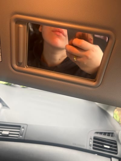 The image displays the interior of a vehicle situated in Lonate Pozzolo, Italy. The upper section of the frame is dominated by a gray-beige sun visor containing an integrated vanity mirror. The mirror reflects a partial view of an individual, specifically their lower face (lips, chin, and upper neck, where a mole or blemish is visible) and a left hand. The individual's lips are natural or lightly colored. Dark hair is visible framing the face. The reflected hand wears a ring with a yellow stone on the ring finger and holds a dark-colored object, consistent with a smartphone, which emits a warm, orange light. The individual appears to be engaging in an action such as taking a self-portrait via the mirror's reflection. Below the sun visor, the vehicle's gray-black dashboard is visible, featuring two horizontal air vents with slats positioned on the left and right sides. Through the top of the windshield, blurred green foliage and a light-colored ground surface are faintly discernible, indicating an outdoor setting.