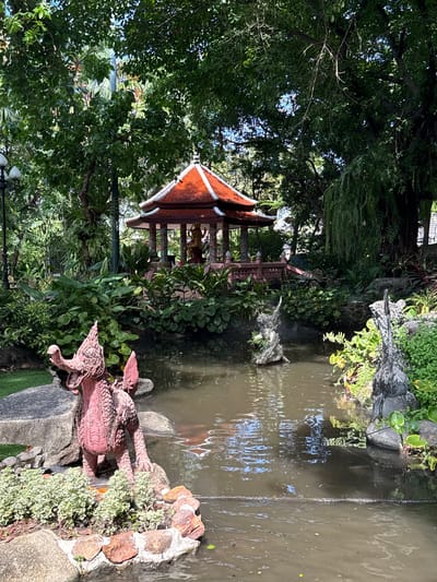 The image shows a pond in a garden setting, located in the Samrong Nuea Subdistrict Municipality, Thailand. The pond's water is murky. At the edge of the pond is a pink dragon statue on a rock formation with a small green grass border. On the opposite side of the pond, there is a gazebo with a red-tiled roof. The scene is surrounded by dense green foliage, with trees and plants obscuring parts of the pond and gazebo. The lighting indicates daylight conditions.
