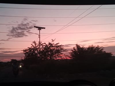 The image shows a dusk or dawn scene in Sagoe, Indonesia, observed from inside a vehicle. The sky is a blend of pink and light purple hues with scattered clouds. A utility pole with a horizontal fixture stands to the left of center. Black electrical wires stretch across the upper portion of the frame. Silhouetted trees and vegetation occupy the middle ground. On the road in the foreground, a motorcycle and a car, both with headlights on, are visible. The lower portion of the image is dark, likely the dashboard of the vehicle.