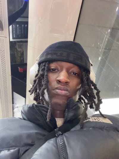 A person with dreadlocks and a black beanie looks directly at the camera. The person wears a dark, puffy jacket. The background appears to be an interior space with white walls and window. The image is rotated to the left. The beanie has some kind of patterns/text on it. The person has dark brown eyes.