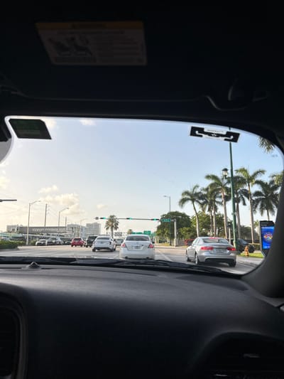 The image was taken from inside a vehicle, looking forward through the windshield at a street scene in Aventura, Florida. The sky is clear with some scattered clouds. Palm trees are visible along the side of the road, and several cars are on the street, either stopped or moving slowly. Traffic lights are visible, with one showing green. Buildings are in the background, including what appears to be a multi-story structure. A billboard is also visible on the right side of the image with text that reads "CELEBRATING 30 YRS". There are stickers on the windshield of the car. The dashboard of the vehicle is in the foreground.