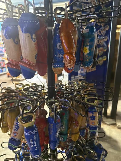 The image displays a close-up of a display rack filled with numerous phallic-shaped bottle opener keychains at a souvenir shop in Ofrynio Beach, Greece. The keychains are made of wood and painted with various designs, including Greek patterns, flowers, evil eyes, and the text "WELCOME TO GREECE." The keychains are arranged in tiers, with the top tier featuring larger, more detailed items and the lower tiers filled with a dense collection of smaller ones. The background is blurred, suggesting an indoor setting with merchandise and lighting typical of a retail establishment.
