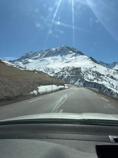 The image depicts a view from a vehicle traveling on a road in Villar-d'Arêne, France. The road appears to be ascending a mountainous area. A prominent, snow-covered mountain range rises to the left of the road. The landscape transitions from barren, grassy slopes near the roadside to increasingly snow-covered terrain higher up the mountain. The road is paved and has white lane markings. Metal guardrails are visible along the edge of the road. The sky is clear and blue with visible sun glare. A portion of the interior of the vehicle is visible on the right side of the frame.