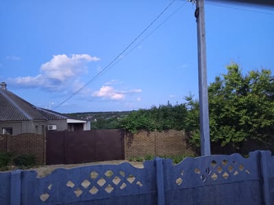 The image depicts an outdoor scene at dusk, likely in Vilshany, Ukraine. The foreground features a blue, decorative concrete fence with a diamond pattern. Behind this, a large, dark brown metal gate is visible, partially obscuring a textured brown brick fence. To the left of the gate is a portion of a grey house with a dark grey, corrugated roof and a chimney. To the right of the gate and behind the brick fence, a large green tree with dense foliage dominates the midground. Further in the distance, a rolling landscape of trees and buildings can be seen under a blue sky with scattered white clouds. A utility pole with wires runs diagonally across the upper portion of the image. The overall impression is of a residential or semi-rural property at the end of the day.