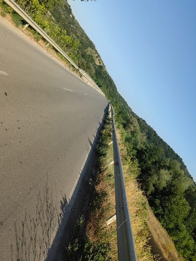 This media file depicts a road with a metal guardrail on the right side, bordered by a wooded hillside. The road appears to be an asphalt surface with white lane markings visible. The hillside is covered in dense green foliage, suggesting a natural, undeveloped area. The sky is a clear, bright blue, indicating a sunny day. The perspective is from a low angle, looking down the road as it curves into the distance. The shadows cast on the road suggest the sun is relatively high in the sky, likely midday or early afternoon. There are no people or vehicles visible in the image. The overall impression is one of a scenic, possibly rural, highway. The scene is set in Staliyska mahala, Bulgaria, a location known for its natural beauty.