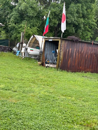 The scene shows a residential area with unkempt vegetation. A wooden shed stands partially in the frame. Adjacent to it, a makeshift structure supports a white boat bearing the number "BC 2362" and a Bulgarian flag. Additional items are haphazardly stacked around the boat, including bottles, buckets, and other indeterminate objects. Two flags, one Japanese and one Bulgarian, are affixed to the structure.
The location is Dabovets, Bulgaria. No people are visible.