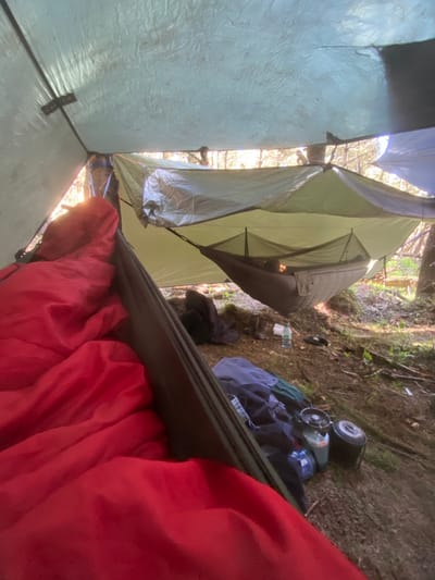 The image shows a view from inside a hammock suspended between two trees, with a tarp overhead providing shelter. The hammock is made of green fabric with a black net inside. It is partially obscured by the tarp. The hammock is occupied by a person.  The individual is wrapped in a red sleeping bag.  The hammock is located under a tarp, strung between trees.  The tarp is a green tarp with a blue and white tarp below it. On the ground are a dark-colored backpack, two water bottles, and a camping stove. The environment is a forest with green vegetation and trees surrounding the campsite. The location is a forested area within the High Peak District of the United Kingdom, based on the provided coordinates.