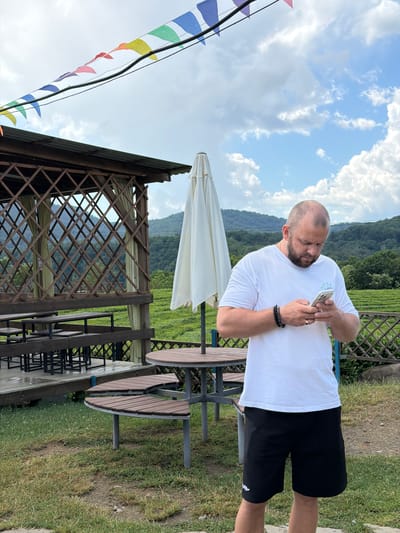 In Izmaylovka, Russia, a bald man with a beard, wearing a white t-shirt and black shorts, stands outside and looks at his phone. He has a bracelet on his left wrist and a ring on his right ring finger. Behind him, a wooden gazebo with tables and benches is visible, along with a large white umbrella and a circular table with seating. The background shows lush green tea plantations stretching towards rolling hills under a partly cloudy sky. Colorful triangular flags are strung across the top of the image.