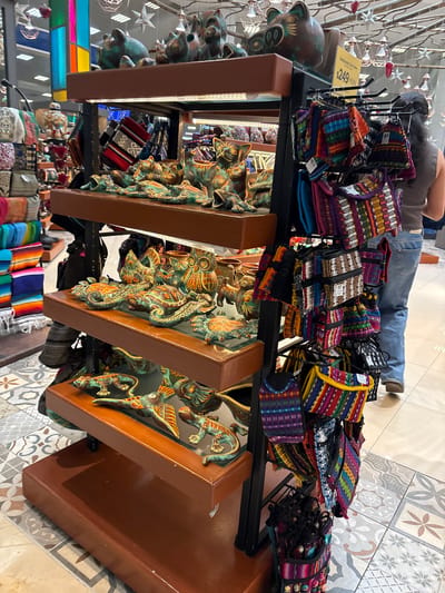 The image depicts a retail display of artisanal crafts in Cancún, Mexico. The display rack features several shelves stocked with colorful woven bags and coin purses, alongside decorative ceramic animal figurines, including cats and turtles, with a distressed green and orange patina. The merchandise is arranged on wooden shelves and hanging from metal racks. In the background, a woman in blue jeans and an orange tank top is browsing the items. Above the display, a yellow sign reads "$249 MXN". The flooring is tiled with a geometric pattern. The overall environment suggests a souvenir shop or a market stall, common in tourist areas of Cancún.