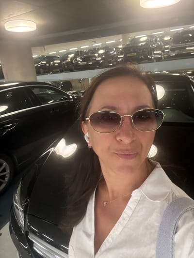 A woman is taking a selfie in a parking garage. She is wearing a white shirt, sunglasses, and earbuds. A black car is visible in the foreground. In the background, other cars are parked on multiple levels. The lighting suggests it is indoors, possibly at night or in an underground garage. The setting appears to be a modern parking facility. There is no specific visual information within the image that allows for identification of the city of Groningen or the Netherlands.