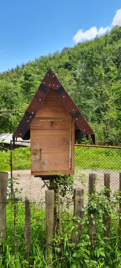 This outdoor image, captured in Iamna, Bulgaria, depicts a small, rustic wooden structure positioned in a verdant, hilly landscape under a clear sky. The central subject is a miniature, house-like wooden box, possibly a mailbox or a large birdhouse, mounted on an unseen post or support. Its pitched, A-frame roof is covered with alternating reddish-brown and dark grey/black asphalt shingles, secured by numerous visible silver-colored fasteners. The body of the structure is constructed from horizontal wooden planks exhibiting distinct grain patterns. On the front panel, a small, round, brass-colored knob or escutcheon is visible near the top left, alongside a dark, aged metal latch or handle that extends vertically downwards.
In the background, a dense forest of various green trees covers a sloping hillside, suggesting a natural, possibly mountainous, environment. Above the treeline, the sky is a vibrant, cloudless blue, with a single, small white cloud visible on the far right.
In the midground and foreground, a weathered chain-link fence runs horizontally across the scene, appearing somewhat rusted in places. Several sturdy, roughly cut wooden fence posts are visible in the immediate foreground, anchoring the chain-link fence. The ground is covered with lush green grass, wildflowers, and various climbing plants or vines that interweave through the chain-link fence and around the wooden posts, creating a natural, overgrown appearance. No people or animals are present in the scene.
