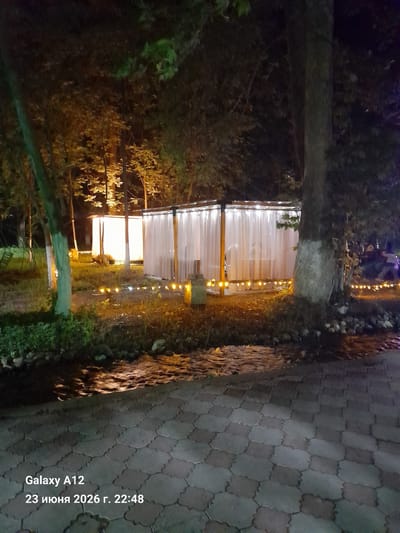 The image depicts a nighttime scene in Kara-Köl City, Kyrgyzstan. The focus is on a structure that appears to be an outdoor seating area or a small pavilion, enclosed by translucent white curtains. Warm yellow lights are strung along the exterior of the structure, creating an inviting ambiance. The area is surrounded by trees and foliage, suggesting a natural or park-like setting. A paved walkway with hexagonal stones is visible in the foreground, partially illuminated by the lights and the dim ambient light of the night. The overall mood is serene and tranquil, enhanced by the soft lighting and the natural elements.
Visible text at the bottom right of the image reads "Galaxy A12 23 июня 2026 г. 22:48", indicating the device used to capture the photo, the date, and the time. The presence of the date "2026" suggests this is a future date. The time is late evening, consistent with the nighttime setting. There are no people visible in the frame, but the setup of the pavilion suggests it might be a place for gatherings or dining. The weather appears calm, with no indicators of rain or wind.