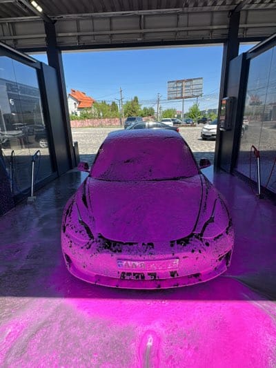 A Tesla car covered in pink foam sits in a car wash bay in Chaiky, Ukraine. The bay is open on one side, revealing a clear, sunny day. In the background, there are several cars parked, a billboard, and buildings. The area around the car is also covered in the pink foam.