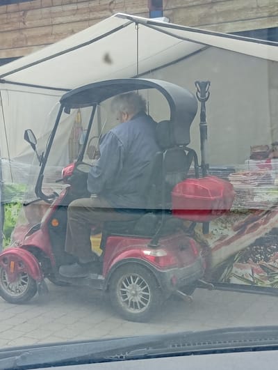 A person is seated on a red three-wheeled scooter, partially obscured by a clear plastic windshield and a black canopy. The person, seen from the back, has gray hair and is wearing a blue shirt and brown pants. A red storage box is attached to the rear of the scooter. The scooter is parked on a paved surface, possibly a sidewalk or market area, with a wooden structure and a white tent in the background. Behind the scooter, there appears to be a display of food items wrapped in plastic. The scene is captured through a car's windshield, indicated by the blurry foreground. The location is identified as Kuršėnai, Lithuania.
